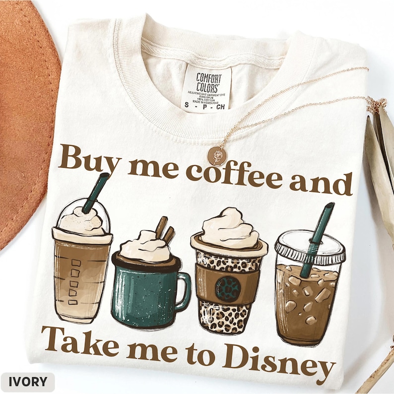 Puede incluir: Camiseta de color marfil con el texto "Buy me coffee and Take me to Disney". La camiseta presenta ilustraciones de cuatro bebidas de caf&eacute; diferentes. Un collar dorado est&aacute; drapeado sobre la camiseta.