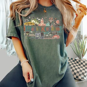 May include: Dark green t-shirt featuring a colorful graphic of a theme park with the text "Magic Kingdom". The design includes illustrations of a castle, rides, and other attractions. The shirt has short sleeves and a relaxed fit.