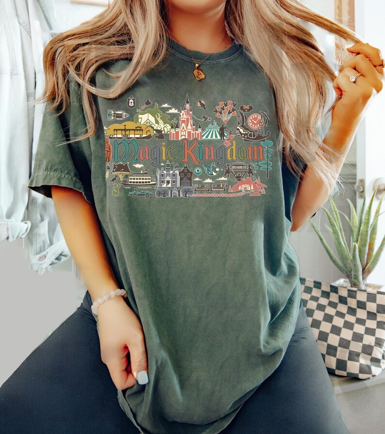 May include: Dark green t-shirt featuring a colorful graphic of a theme park with the text "Magic Kingdom". The design includes illustrations of a castle, rides, and other attractions. The shirt has short sleeves and a relaxed fit.