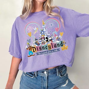 Może przedstawiać: Liliowa koszulka z grafiką Disneyland. Projekt zawiera logo Disneyland, kultowe postacie, takie jak Myszka Miki i Myszka Minnie, oraz zamek. Tekst "The happiest place on earth" i "EST. 1955" są r&oacute;wnież obecne.