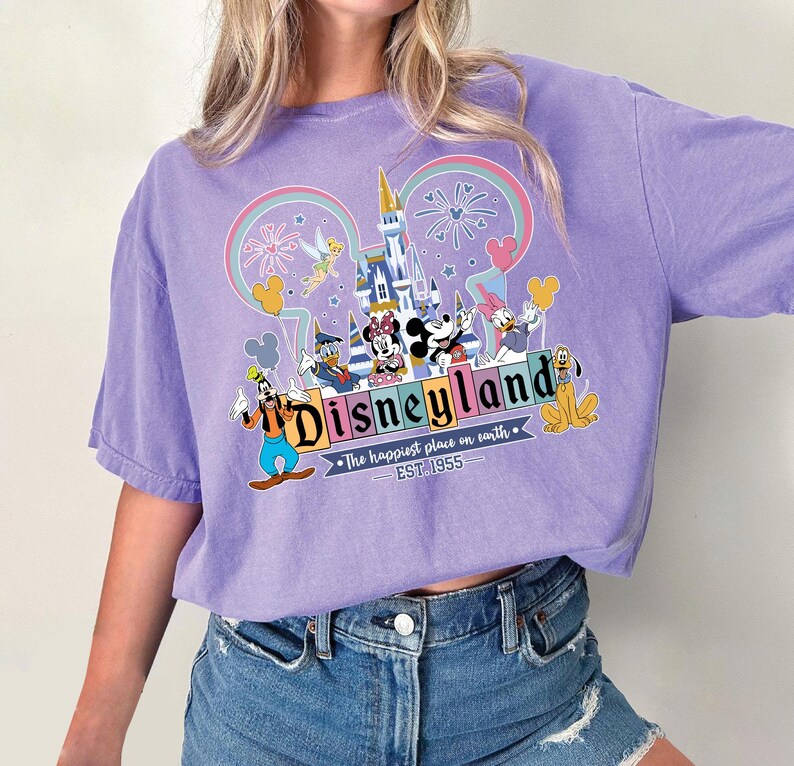 Może przedstawiać: Liliowa koszulka z grafiką Disneyland. Projekt zawiera logo Disneyland, kultowe postacie, takie jak Myszka Miki i Myszka Minnie, oraz zamek. Tekst "The happiest place on earth" i "EST. 1955" są r&oacute;wnież obecne.