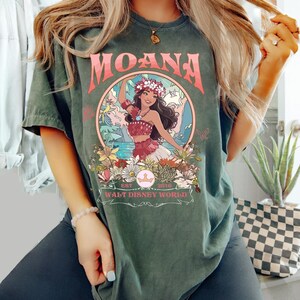 May include: Dark green t-shirt featuring the word "MOANA" in pink lettering above a circular graphic. The graphic depicts a woman in a floral crown and traditional attire, surrounded by flowers and the text "WALT DISNEY WORLD".