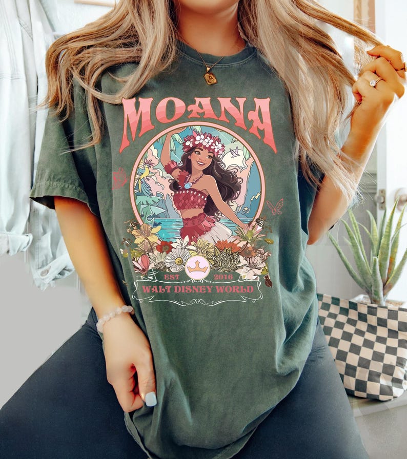 May include: Dark green t-shirt featuring the word "MOANA" in pink lettering above a circular graphic. The graphic depicts a woman in a floral crown and traditional attire, surrounded by flowers and the text "WALT DISNEY WORLD".