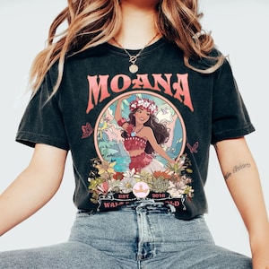 May include: Black t-shirt featuring the word "MOANA" in pink above a circular graphic. The graphic depicts a woman in a floral crown and red outfit, surrounded by flowers and butterflies. The text "EST 2016" and "WALT WORLD" are also visible.