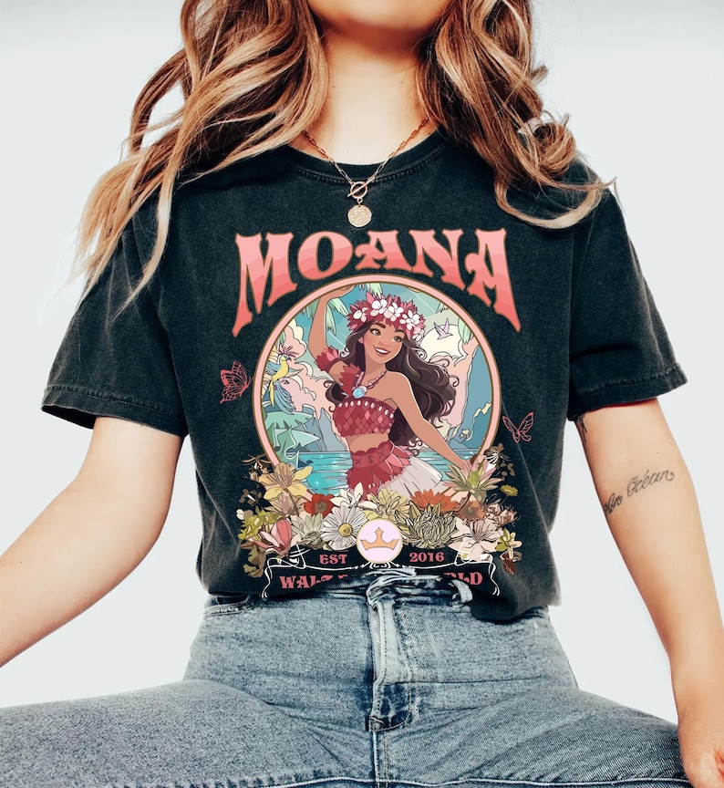 May include: Black t-shirt featuring the word "MOANA" in pink above a circular graphic. The graphic depicts a woman in a floral crown and red outfit, surrounded by flowers and butterflies. The text "EST 2016" and "WALT WORLD" are also visible.