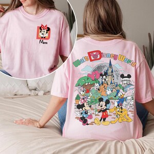 May include: A light pink t-shirt featuring a Minnie Mouse graphic with the word "Mom" in black. The back of the shirt has a colorful graphic of Disney characters and the words "Walt Disney World".