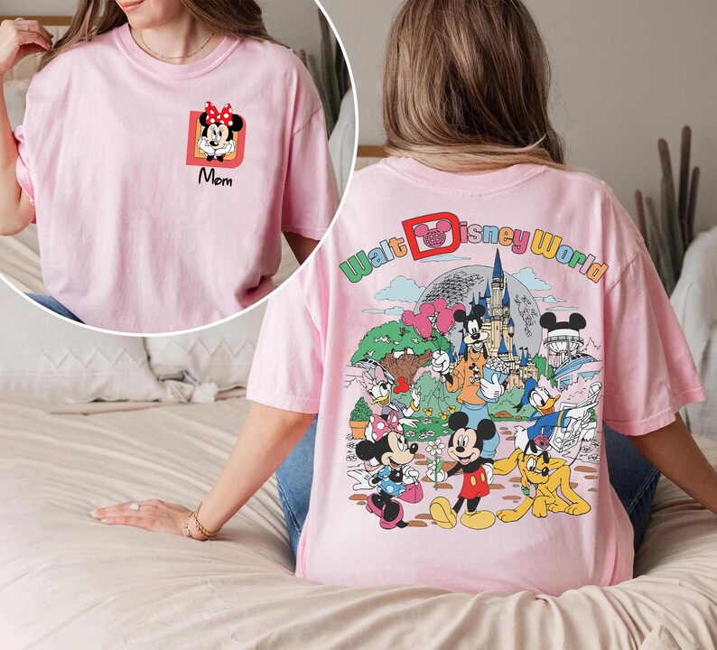 May include: A light pink t-shirt featuring a Minnie Mouse graphic with the word "Mom" in black. The back of the shirt has a colorful graphic of Disney characters and the words "Walt Disney World".
