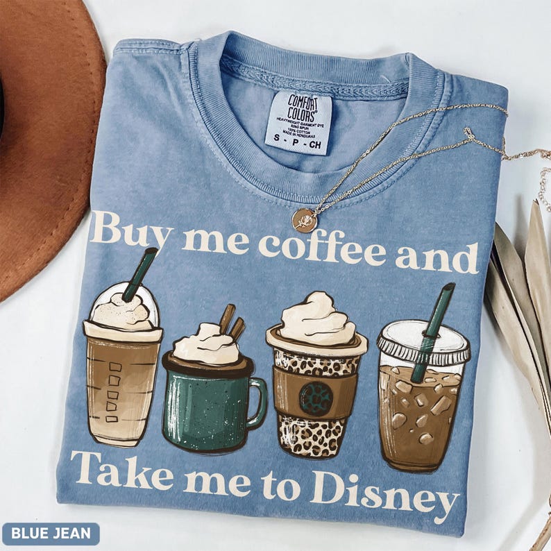 Puede incluir: Camiseta azul vaquero Comfort Colors con el texto "Buy me coffee and Take me to Disney". El gr&aacute;fico presenta cuatro bebidas de caf&eacute;. Un collar dorado est&aacute; drapeado sobre la camisa.