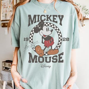May include: A light green t-shirt featuring a distressed graphic of Mickey Mouse. The design includes the words "Mickey Mouse" and "Disney", along with the years "1928" and "19". The shirt has short sleeves and a crew neck.