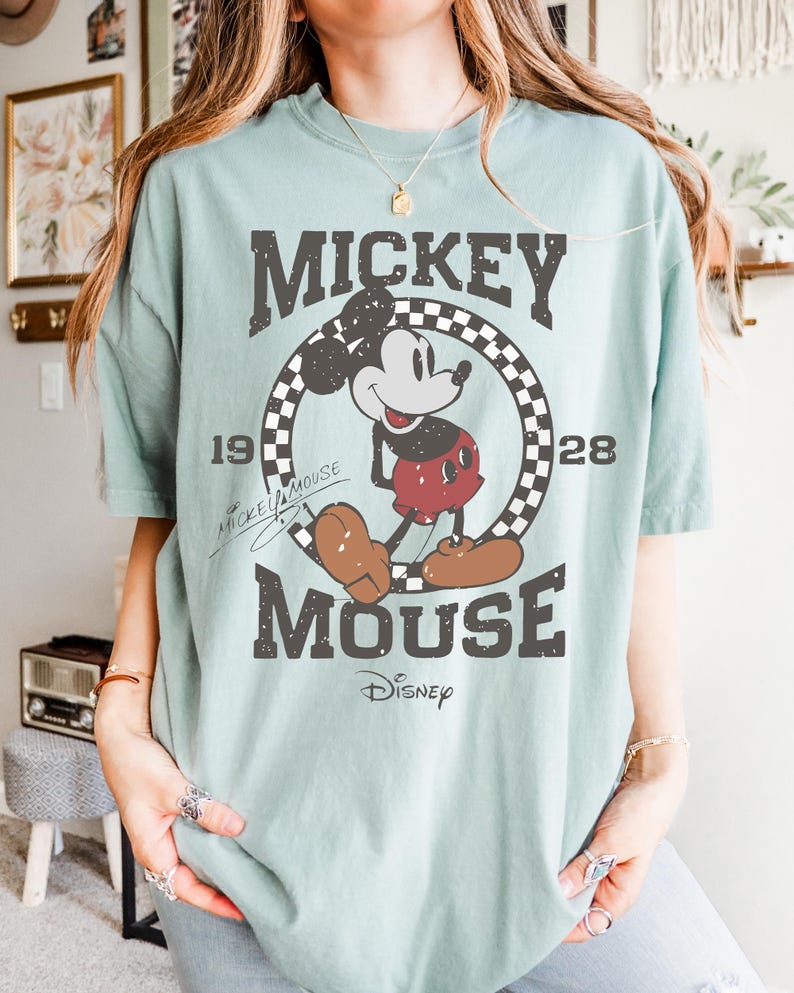May include: A light green t-shirt featuring a distressed graphic of Mickey Mouse. The design includes the words "Mickey Mouse" and "Disney", along with the years "1928" and "19". The shirt has short sleeves and a crew neck.