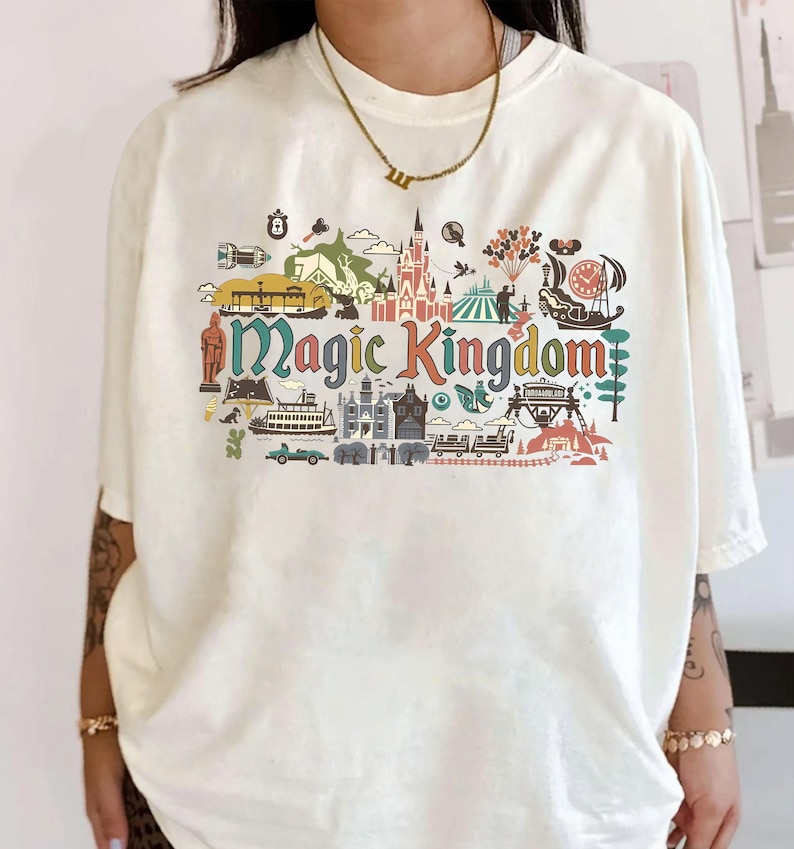May include: Cream-colored t-shirt featuring a colorful graphic of the Magic Kingdom. The design includes various park icons and the words "Magic Kingdom" in a stylized font. The shirt has short sleeves and a relaxed fit.