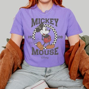 May include: A purple t-shirt featuring a distressed graphic of Mickey Mouse. The design includes the text "Mickey Mouse" in a vintage font, along with the year "1928" and a checkered circle around Mickey. The Disney logo is at the bottom.