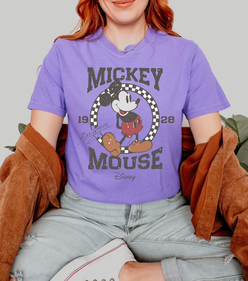May include: A purple t-shirt featuring a distressed graphic of Mickey Mouse. The design includes the text "Mickey Mouse" in a vintage font, along with the year "1928" and a checkered circle around Mickey. The Disney logo is at the bottom.