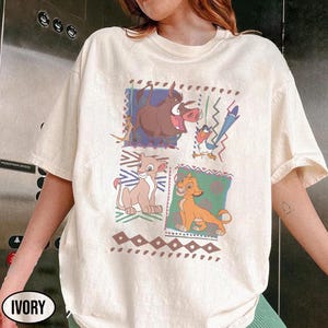 May include: Ivory-colored t-shirt featuring a colorful graphic of characters from The Lion King. The design includes Timon, Pumbaa, Simba, and Nala, set against geometric and patterned backgrounds. The shirt has a classic crew neck and short sleeves.
