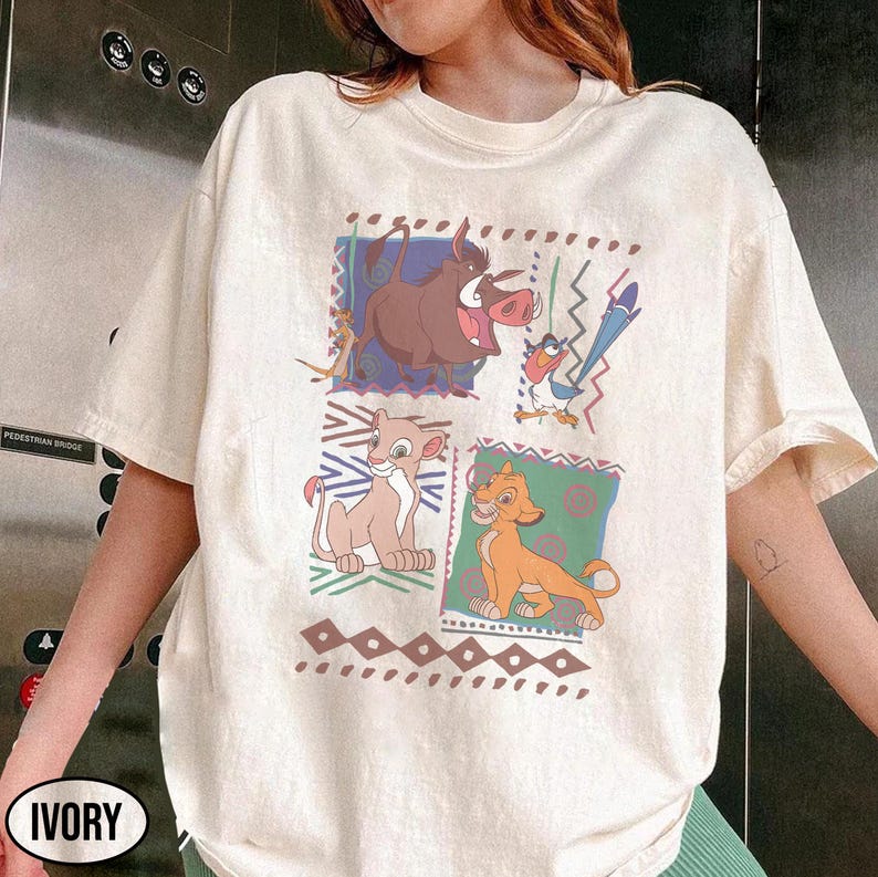 May include: Ivory-colored t-shirt featuring a colorful graphic of characters from The Lion King. The design includes Timon, Pumbaa, Simba, and Nala, set against geometric and patterned backgrounds. The shirt has a classic crew neck and short sleeves.