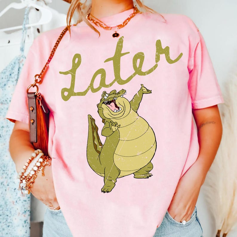 May include: Pink t-shirt with a cartoon alligator graphic and the word "Later" in a cursive font. The alligator is green and yellow, with its mouth open and one arm raised. The shirt is a solid pink color.