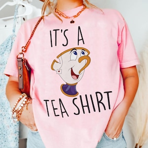 May include: Pink t-shirt with the text "IT'S A TEA SHIRT" and a cartoon image of a teacup. The teacup is white with a gold handle and rim, a purple base, and a small red heart.