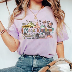 May include: A lilac-colored t-shirt with the words "Magic Kingdom" in a colorful, illustrated design. The graphic includes various theme park elements such as a castle, balloons, and a carousel. The shirt is paired with blue jeans.