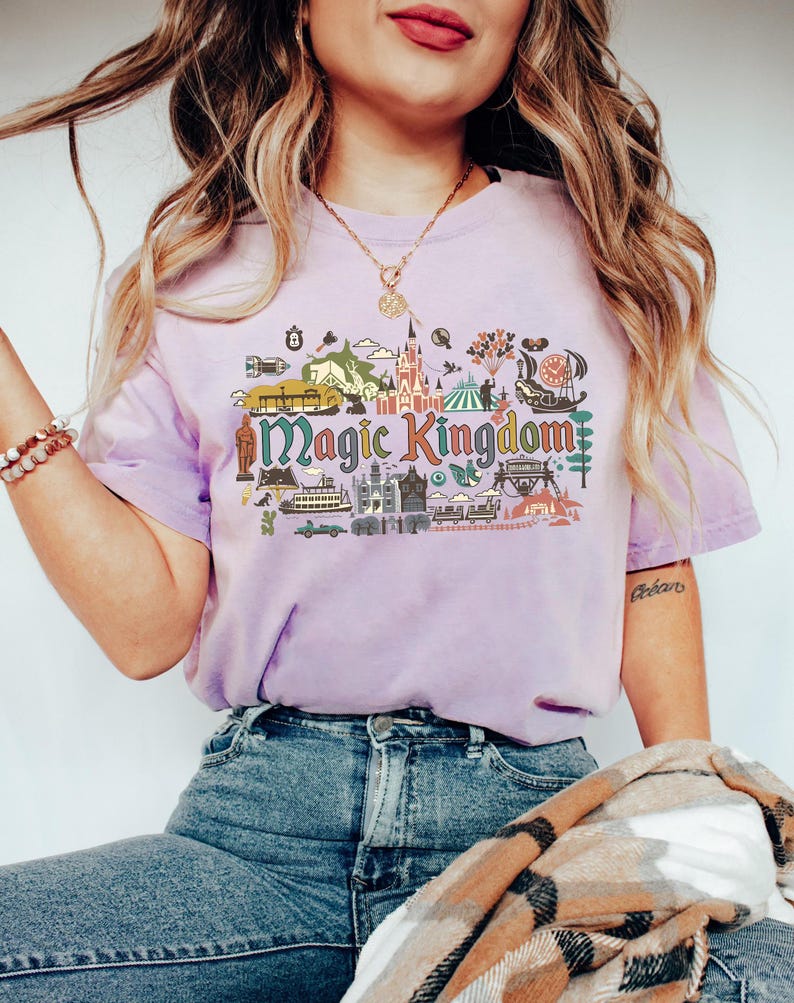 May include: A lilac-colored t-shirt with the words "Magic Kingdom" in a colorful, illustrated design. The graphic includes various theme park elements such as a castle, balloons, and a carousel. The shirt is paired with blue jeans.