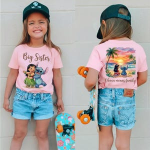 May include: Two young girls wearing matching pink t-shirts and denim shorts. The front of the shirts say "Big Sister" and feature a graphic of Lilo and Stitch. The back of the shirts have a beach scene with the words "Ohana means family".