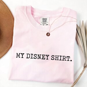 May include: A light pink t-shirt with the text "MY DISNEY SHIRT." printed in black. A silver necklace with a circular pendant rests on the shirt. The shirt is folded and has a tag that says "COMFORT COLORS."