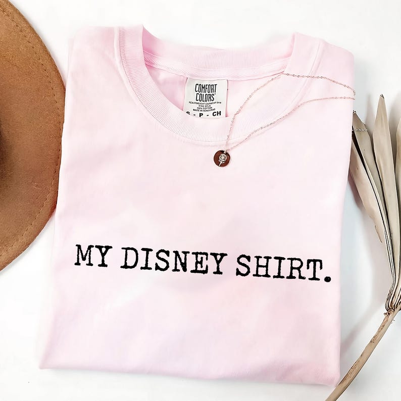May include: A light pink t-shirt with the text "MY DISNEY SHIRT." printed in black. A silver necklace with a circular pendant rests on the shirt. The shirt is folded and has a tag that says "COMFORT COLORS."