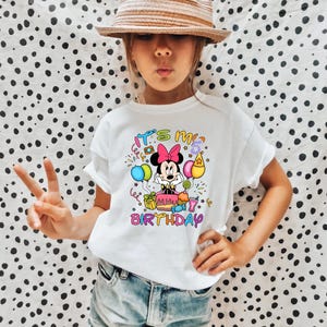 May include: White t-shirt featuring a colorful graphic of Minnie Mouse with balloons, a birthday cake, and the text "It's my 5th Birthday". The design includes pink, blue, yellow, and green elements.