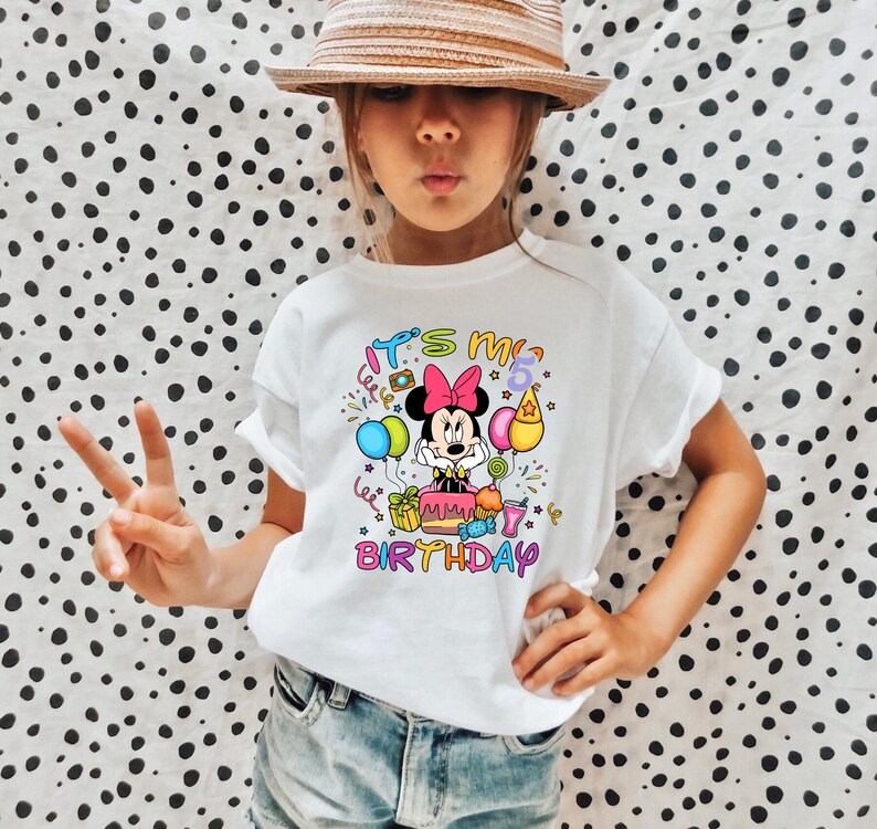 May include: White t-shirt featuring a colorful graphic of Minnie Mouse with balloons, a birthday cake, and the text "It's my 5th Birthday". The design includes pink, blue, yellow, and green elements.