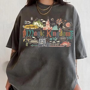 May include: Dark gray t-shirt featuring a colorful graphic of the Magic Kingdom. The design includes various park icons and the words "Magic Kingdom" in a stylized font. The shirt has short sleeves and a relaxed fit.