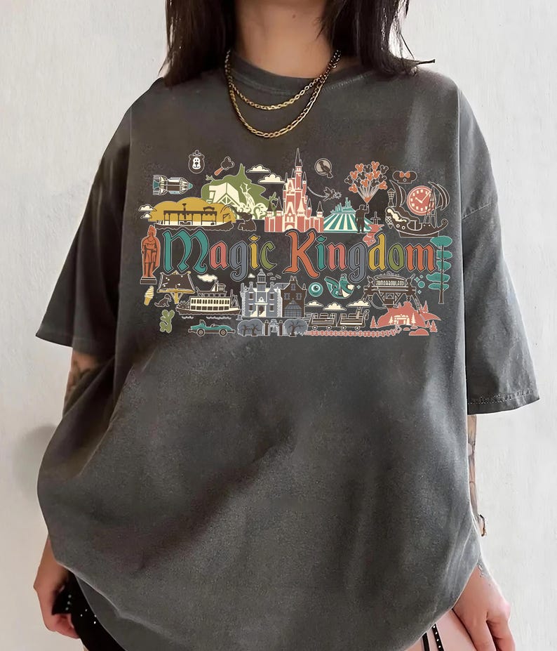 May include: Dark gray t-shirt featuring a colorful graphic of the Magic Kingdom. The design includes various park icons and the words "Magic Kingdom" in a stylized font. The shirt has short sleeves and a relaxed fit.