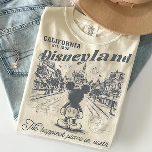 May include: Cream-colored t-shirt with a Disneyland graphic. The design features Mickey Mouse, a street scene, and the text "Disneyland California Est. 1955" and "The happiest place on earth". The shirt is folded on top of blue jeans.