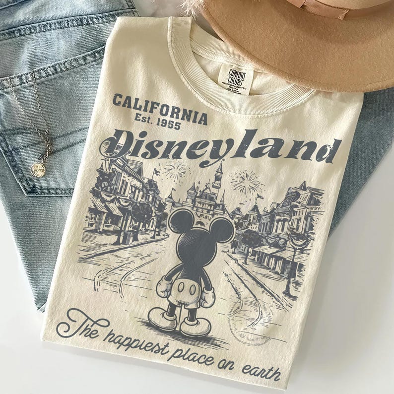 May include: Cream-colored t-shirt with a Disneyland graphic. The design features Mickey Mouse, a street scene, and the text "Disneyland California Est. 1955" and "The happiest place on earth". The shirt is folded on top of blue jeans.