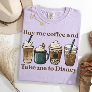 Puede incluir: Una camiseta lila con el texto "Buy me coffee and Take me to Disney" e ilustraciones de cuatro bebidas de caf&eacute; diferentes. Se ve un collar dorado y un sombrero.