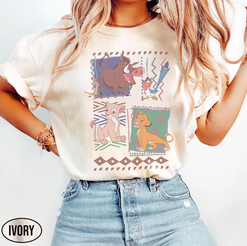 May include: Ivory-colored t-shirt featuring a colorful graphic of characters from The Lion King. The design includes images of Pumbaa, Timon, Simba, and Nala, set against geometric backgrounds. The word "IVORY" is printed at the bottom.