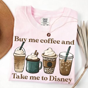 Puede incluir: Una camiseta rosa claro con el texto "Buy me coffee and Take me to Disney" en marr&oacute;n. La camiseta presenta ilustraciones de cuatro bebidas de caf&eacute; diferentes. Un collar plateado con un peque&ntilde;o colgante est&aacute; drapeado sobre la camiseta.