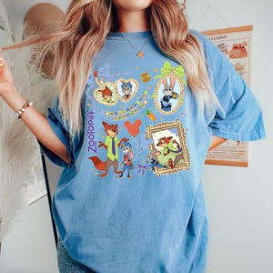 May include: A light blue t-shirt featuring colorful cartoon characters from the movie Zootopia. The shirt has a crew neck and short sleeves. The graphic includes Nick Wilde, Judy Hopps, and other characters in framed designs. The word "Zootopia" is printed vertically on the left side.