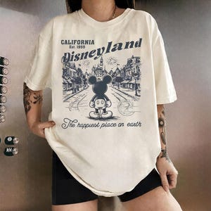 May include: Cream-colored t-shirt featuring a Disneyland graphic. The design includes an illustration of Mickey Mouse, the text "Disneyland California Est. 1955," and the phrase "The happiest place on earth."