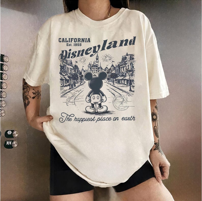 May include: Cream-colored t-shirt featuring a Disneyland graphic. The design includes an illustration of Mickey Mouse, the text "Disneyland California Est. 1955," and the phrase "The happiest place on earth."