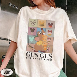 Peut inclure: T-shirt ivoire avec une illustration de Gus Gus de Cendrillon tenant des collations. Le design comprend une grille d'icônes alimentaires et le texte "GUS GUS THE SNACK TOUR".