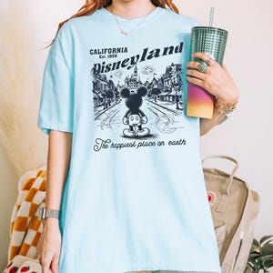May include: Light blue t-shirt featuring a black and white graphic of Disneyland with Mickey Mouse. The text on the shirt reads "CALIFORNIA Disneyland Est. 1955" and "The happiest place on earth."