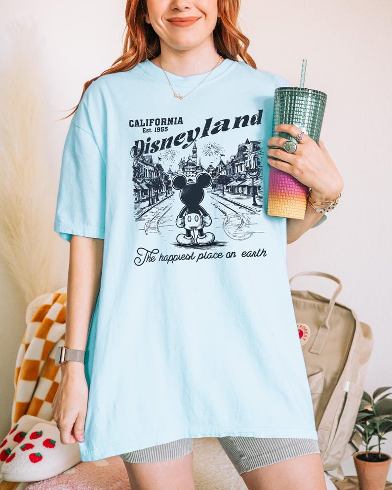 May include: Light blue t-shirt featuring a black and white graphic of Disneyland with Mickey Mouse. The text on the shirt reads "CALIFORNIA Disneyland Est. 1955" and "The happiest place on earth."