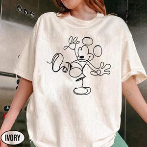 May include: Ivory-colored t-shirt featuring a black line drawing of Mickey Mouse. The design includes the character's iconic ears, face, and body in a minimalist style. The word "Ivory" is printed on the bottom left of the shirt.