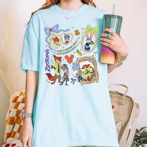 May include: Light blue t-shirt featuring a colorful graphic of characters from the animated film "Zootopia." The design includes framed portraits, character illustrations, and the film's title. The shirt is a comfortable, casual style.