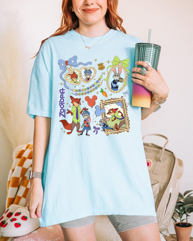 May include: Light blue t-shirt featuring a colorful graphic of characters from the animated film "Zootopia." The design includes framed portraits, character illustrations, and the film's title. The shirt is a comfortable, casual style.