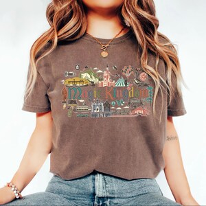 May include: A brown t-shirt with a colorful graphic design featuring the words "Magic Kingdom" and various illustrations of theme park attractions. The shirt has short sleeves and a crew neck.