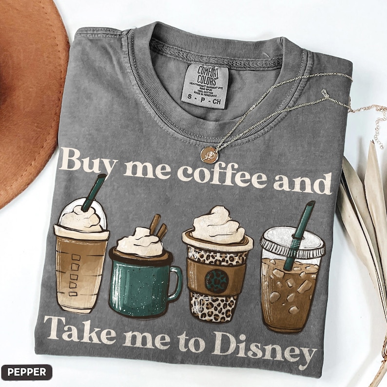 Puede incluir: Camiseta gris Comfort Colors con el texto "Buy me coffee and Take me to Disney". La camiseta presenta ilustraciones de cuatro bebidas de caf&eacute; diferentes. Un collar plateado con un peque&ntilde;o colgante est&aacute; sobre la camiseta.