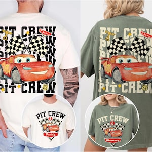 May include: Two t-shirts, one cream and one olive green, with a cartoon race car design. The design includes the text "PIT CREW", checkered flags, and a Route 66 sign. The cream shirt has a smaller design that says "Daddy". The olive green shirt has a smaller design that says "Mommy".