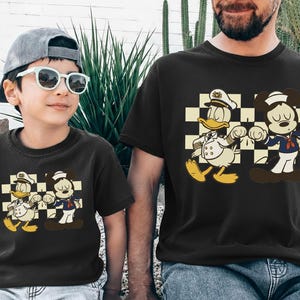 May include: Black t-shirts featuring a cartoon graphic of Donald Duck and Mickey Mouse in sailor outfits. The characters are set against a checkered background. The shirts are worn by a child and an adult.