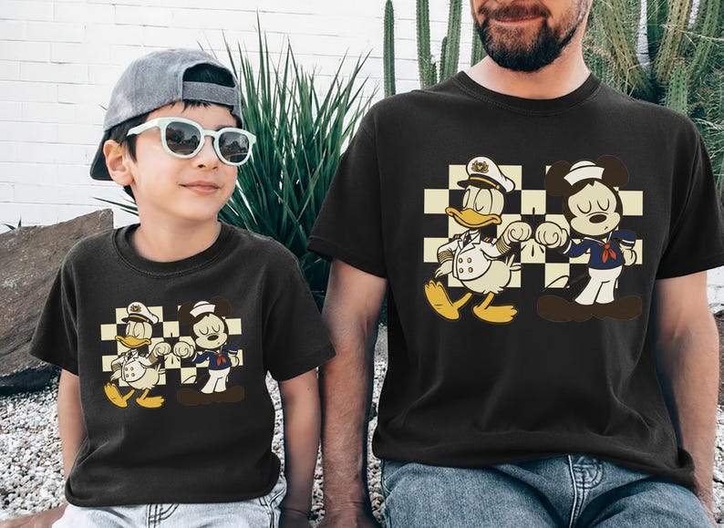 May include: Black t-shirts featuring a cartoon graphic of Donald Duck and Mickey Mouse in sailor outfits. The characters are set against a checkered background. The shirts are worn by a child and an adult.