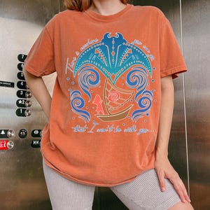 May include: Orange t-shirt with a graphic design featuring a boat, waves, and the text "There is nowhere you can go that I won't be with you..." The design is in shades of blue, orange, and brown. The shirt is worn with striped shorts.